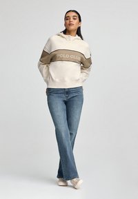 Felpa crema con una striscia marrone che presenta la scritta "POLO CLUB" in beige, abbinata a jeans a zampa d'elefante blu e sneaker beige. Tessuto di qualità liscia.