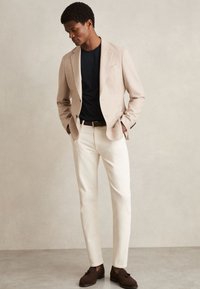 Mann trägt beigen Blazer, schwarzes Hemd, weiße Hose und braune Loafers, steht mit den Händen in den Taschen vor neutralem Hintergrund.