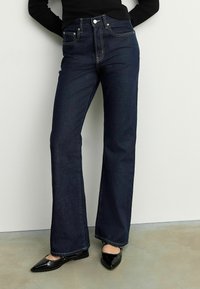 Jean taille haute bleu foncé en denim, avec une coupe droite, des coutures contrastantes et des poches avant, associés à des chaussures noires.