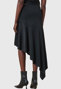 Femme portant une jupe noire asymétrique avec un ourlet haut-bas et des bottes noires à talons mi-mollet, vue de dos sur un fond gris.