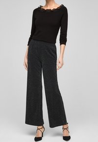 Kvinna i en svart långärmad topp med volangkrage och svarta glittriga vida byxor, i kombination med svarta remsandaler.