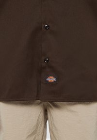 Detailní záběr na hnědou košili s knoflíky s logem Dickies nad pasem béžových kalhot.