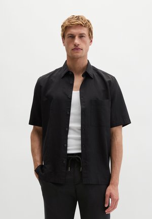 Blonde man draagt een zwart kort mouw overhemd met knopen over een wit tanktop, met een zwarte broek, staand tegen een effen lichte achtergrond.