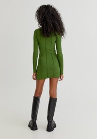 Groene geribbelde lange jurk met een aansluitend ontwerp en een rits aan de achterkant. Gepaarde met kniehoge zwarte rubberen laarzen met een dikke zool.