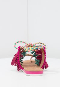 Sandale à bout ouvert avec une semelle rose, une semelle intérieure en cuir brun rembourrée et une sangle tressée multicolore. Présente des pompons en vert foncé et des accents roses.