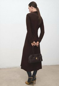 Vestido marrón de manga larga, con corsé ajustado y falda fluida. Bolso oscuro con herrajes dorados, combinado con calcetines azules y zapatos de tacón color coñac.