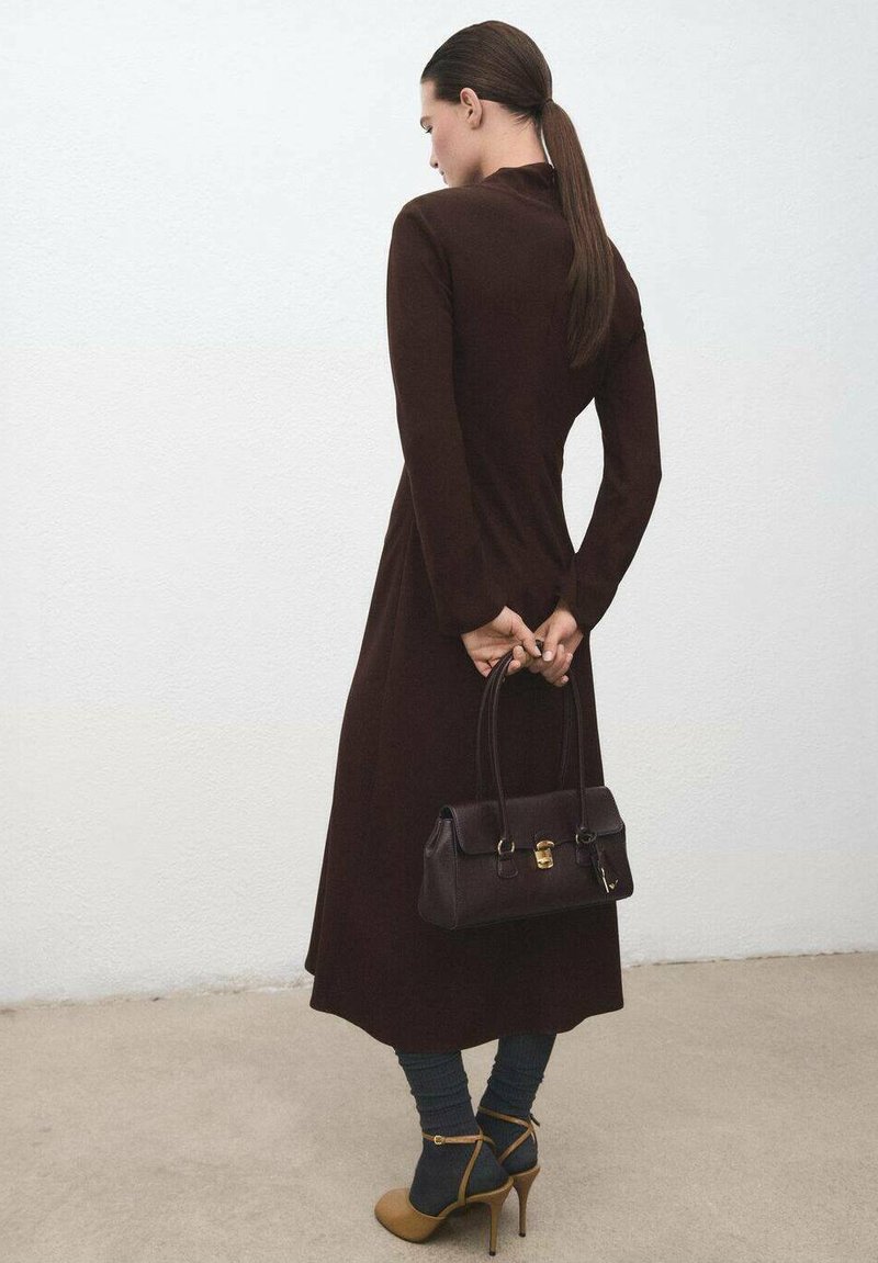 Vestido marrón de manga larga, con corsé ajustado y falda fluida. Bolso oscuro con herrajes dorados, combinado con calcetines azules y zapatos de tacón color coñac.