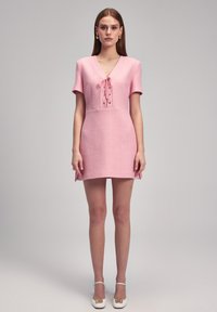 Kurzes pinkes Kleid mit kurzen Ärmeln, V-Ausschnitt, Schnürdetail, strukturiertem Stoff und seitlichen Schlitzen, kombiniert mit weißen Absatzschuhen.