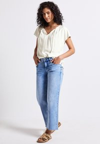 Top corto de color crema claro con manga corta y escote en V, combinado con jeans de cintura alta azul claro y sandalias deslizantes color beige. Atuendo casual con un ajuste relajado.