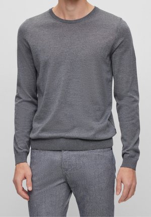 Mann trägt einen enganliegenden grauen Langarm-Pullover mit Rundhalsausschnitt und passende graue Hose, steht vor schlichtem Hintergrund.