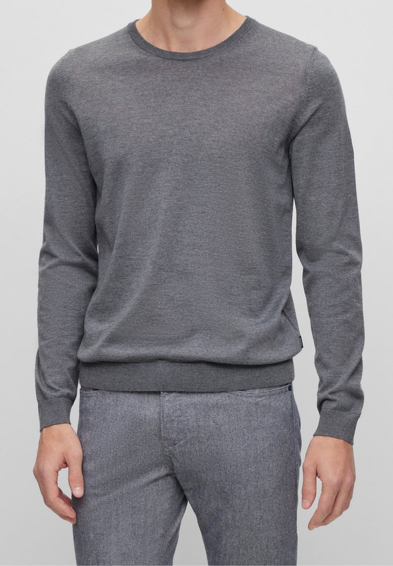 Homme portant un pull gris ajusté à manches longues et col rond avec un pantalon gris assorti, debout devant un fond uni.