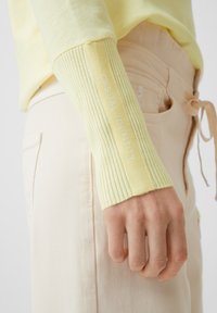 Gul stickad tröja med ribbade ärmmuddar. Ärmmudden har texten "CASUAL IDENTITY" i vitt. Kombinerad med ljusbeige byxor.