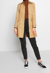 Beige kevyt trench coat, jossa on kaulus, napitettu etuosa, yhdistetty ruskeaan ribattu villapaitaan ja mustiin korkeavyötäröisiin farkkuihin sekä mustiin lenkkareihin.