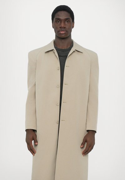 Beige overcoat with a relaxed fit, large notched collar, six buttons, and minimal detailing, worn over a dark shirt. Smooth texture.
