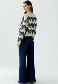 Strickpullover in Creme mit schwarzen Fransen; weite blaue Jeans; glatte Textur; lässige Passform; runder Halsausschnitt.