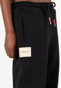 Pantalon de survêtement noir en tissu doux avec une taille à cordon, des poches latérales, et une étiquette crème indiquant « STUDIO EDITION » sur la cuisse.