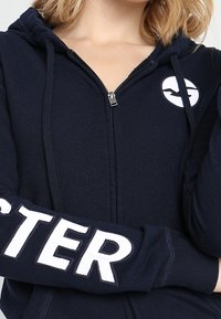 Felpa con zip blu navy, dotata di un logo rotondo con un uccello, grande testo bianco "HUSTLER" sulla manica sinistra, e un tessuto morbido e strutturato.