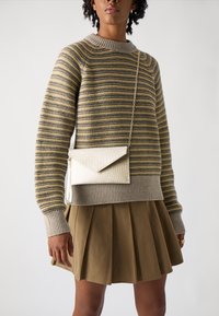 Beige gestreepte trui met geribbelde manchetten en onderrand, gecombineerd met een geplooide khaki rok. Een kleine, metalen envelopvormige tas hangt aan een ketting.