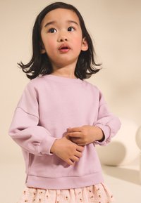 Vaaleanpunainen crew-neck collegepaita, jossa on puffatut hihat ja ribbireunat, yhdistettynä kuviolliseen vaaleanpunaiseen hameeseen, jossa on pieniä koristeellisia yksityiskohtia.