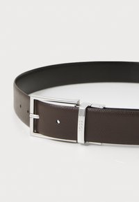 Ceinture en cuir marron avec une finition texturée, équipée d'une boucle en métal argenté et d'un logo. Des trous pour des ajustements de taille le long de la sangle.