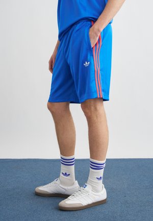 Άνδρας που φοράει μπλε σορτς Adidas με κόκκινες ρίγες, λευκές κάλτσες Adidas με μπλε ρίγες και λευκά αθλητικά παπούτσια Adidas, στέκεται σε μπλε δάπεδο.