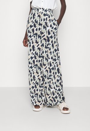 Kvinde iført højttaljede, vide bukser med blå, sort og beige dyreprint, hvid top og hvide åbne sandaler på en ensfarvet baggrund.