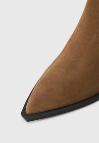 Bottine en daim marron avec un bout pointu et une semelle en caoutchouc noir. Texture lisse avec des lignes épurées et sans embellissements visibles.