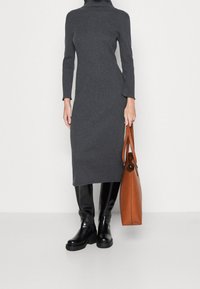 Donna che indossa un abito midi a collo alto grigio scuro attillato, stivali neri fino al ginocchio, mentre tiene una grande borsa a tracolla in pelle marrone.