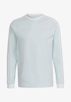 Homme portant un haut à manches longues Adidas bleu clair avec des rayures blanches et un pantalon cargo gris Adidas, debout avec une main dans la poche.