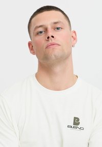 Fehér pamut póló, sötétzöld logóval a bal mellkasi részen. A modellen rövid haj van, és közvetlenül a kamerába néz.