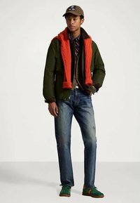 Joven con una gorra marrón, chaqueta verde, suéter naranja sobre los hombros, vaqueros azules y zapatillas verdes, de pie sobre un fondo claro.