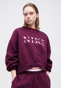 Giovane che indossa un coordinato di felpa e pantaloni Nike viola, con le mani in tasca, che guarda direttamente avanti su uno sfondo semplice.