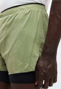 Χακί αθλητικό σορτς από υφή υφάσματος, με ελαστική μέση και μικρό λογότυπο. Φαίνεται μαύρη εσωτερική επένδυση.