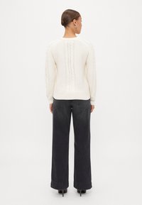 Maglione spesso a maglia crema con orlo a coste, caratterizzato da un motivo a treccia verticale lungo il retro; abbinato a jeans neri a gamba larga.