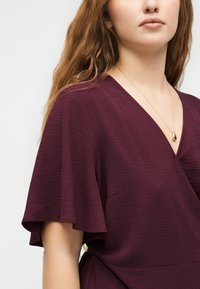 Γυναίκα με μακριά ανοιχτό καστανά μαλλιά φοράει μια υφασμάτινη φούξια μπλούζα με V-λαιμόκοψη και φουσκωτά μανίκια, και ένα χρυσό κολιέ με κρεμαστό δάκρυ.