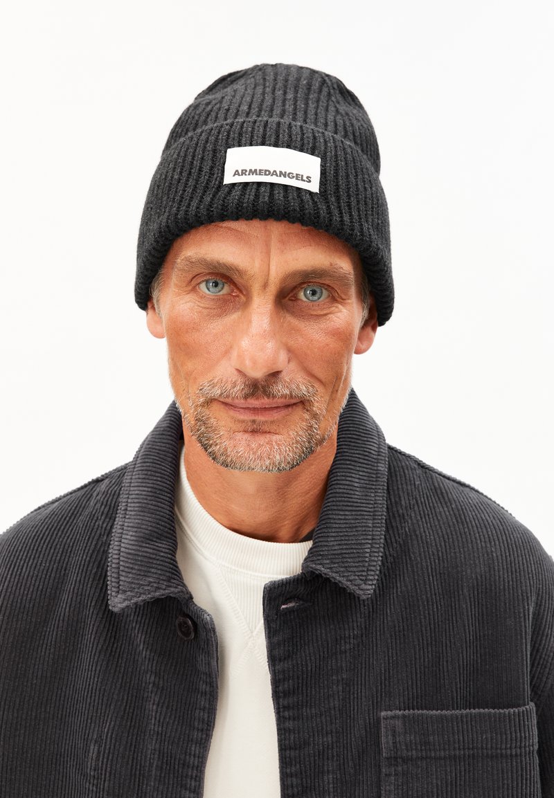 Bonnet en tricot noir à côtes avec une étiquette rectangulaire blanche portant l'inscription "ARMEDANGELS". L'arrière-plan est blanc uni, mettant en valeur la texture du bonnet.