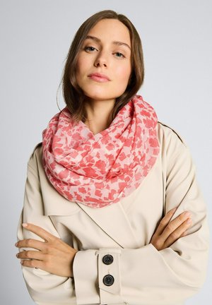 Jonge vrouw met bruin haar, gekleed in een beige trenchcoat en een oneindige sjaal met roze bloemmotief, armen gekruist tegen een effen achtergrond.