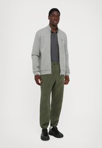 Mann som står med en lys grå jakke med glidelås, mørk grå polo skjorte, olivengrønne teksturerte bukser og svarte joggesko mot en ensfarget bakgrunn.