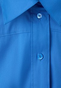 Chemise à col bleu vue de face avec deux boutons visibles et coutures détaillées sur un tissu lisse.