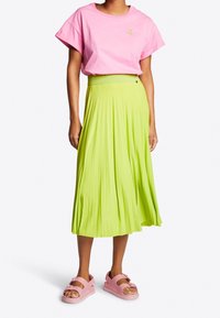 Person klädd i en rosa kortärmad topp, limegrön veckad midikjol och rosa plattformssandaler som står mot en vit bakgrund.