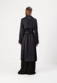 Cappotto nero lungo con vita regolabile tramite cintura, spalline e dettagli plissettati sul retro. Tessuto liscio e design fino al ginocchio.