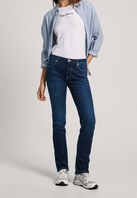 Mörkblå jeans med slim fit, kompletterat med en vit t-shirt med en logotyp. En ljusblå randig skjorta är lagd över outfiten.