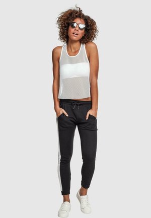 Top sin mangas de malla blanca sobre un sujetador blanco, combinado con joggers negros con rayas laterales blancas y zapatillas blancas. Lleva puestas unas gafas de sol plateadas.