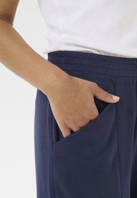 Námořnické modré elastické kalhoty s měkkou texturou, se bočními kapsami; ruka modelky je umístěna v levé kapse, částečně viditelná.