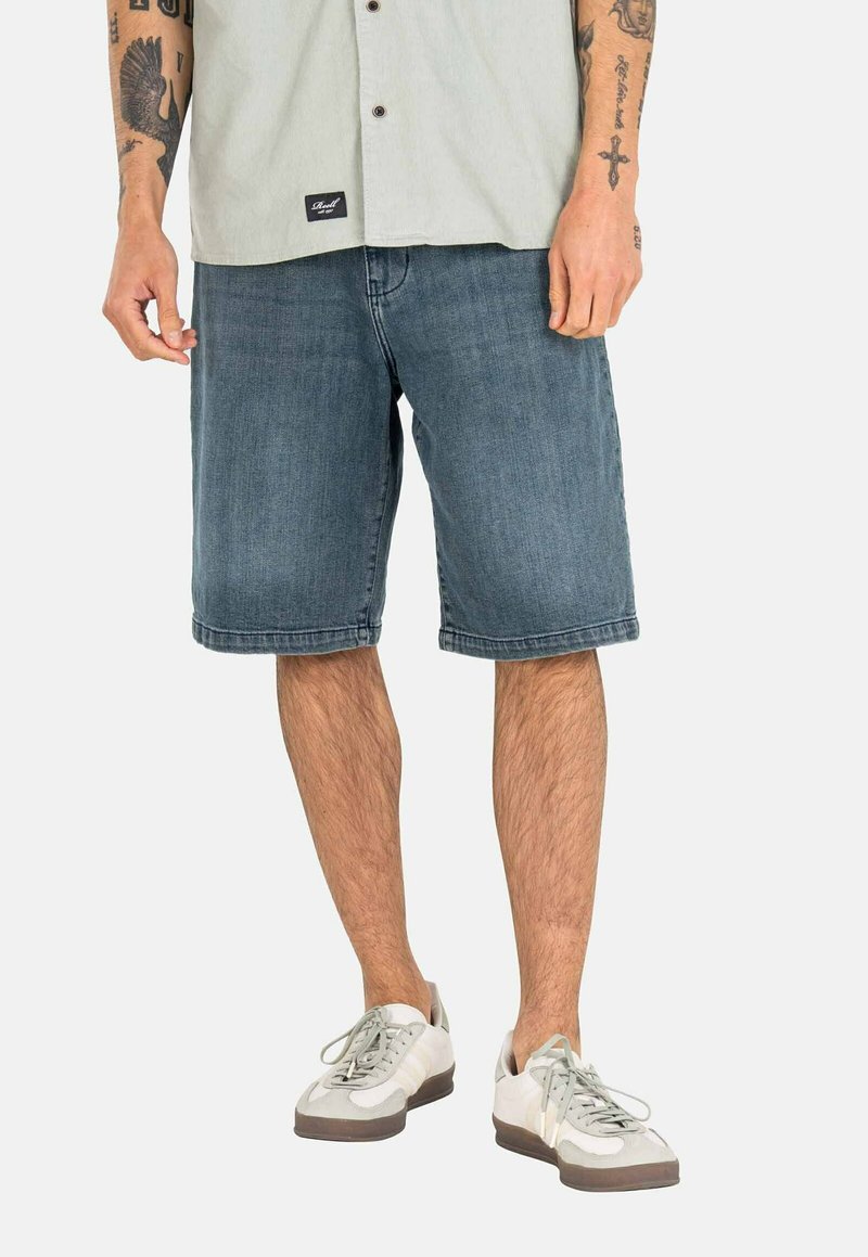 Pantalones cortos de mezclilla en un azul desgastado, hasta la rodilla y con un corte relajado. Combinados con una camisa de botones en gris claro y zapatillas ligeras.