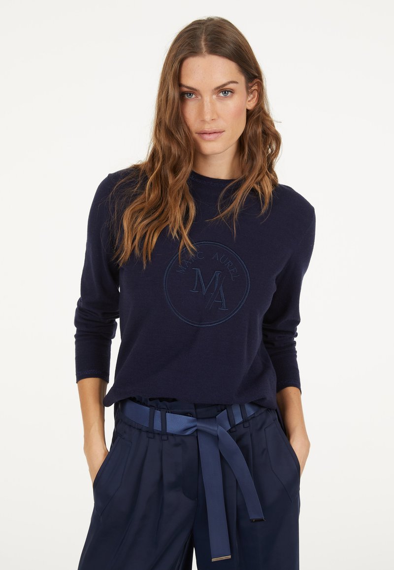 Navy blue sweater with long sleeves and round neckline. Features a tonal embroidered logo at the chest. Paired with dark trousers and belt.