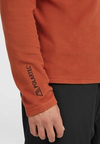 Orange fleece långärmad topp med en mjuk textur. Har en svart "Polartec"-logotyp på ärmslutet och en rak passform.