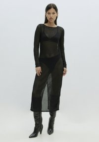 Schwarzes, langärmeliges, durchsichtiges Strickkleid mit einer figurschmeichelnden Silhouette, ausgestattet mit einem seitlichen Schlitz und kombiniert mit knielangen, spitzen Stiefeln.