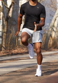 Czarna koszulka sportowa, jasnoszare szorty, białe buty do biegania. Skoncentruj się na postaci mężczyzny w ruchu na ścieżce wśród drzew z nagimi gałęziami.