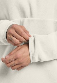 Felpa bianca in fleece con superficie testurizzata; polsini elasticizzati con una fascia interna a contrasto; primo piano di mani che sistemano la manica.